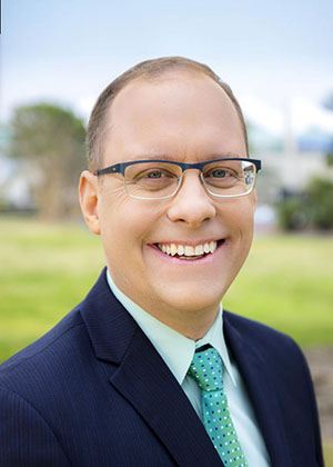 Mr. Jason Giffen, looking clean-cut and business-like in a blue suit and tie against a green lawn background, he is smiling and wearing glasses 