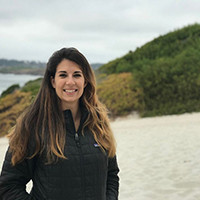 Kelly Leo, Director of the Resilient Coasts Program for The Nature Conservancy in Maryland and Washington D.C., she is standing on a white sand beach with green dunes and a bay behind her, she's wearing a black warm jacket, long brown hair around her shoulders, and a warm smile