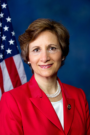 The Honorable Suzanne Bonamici, Congresswoman, First Congressional District of Oregon wearing a red blazer against a blue background and US flag, looking smart and determined