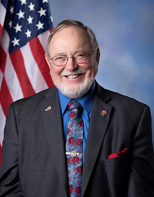 The Honorable Don Young, Congressman, Alaska’s only Representative to the United States House of Representatives, wearing a gray blazer, with blue and red tie, US flag in background, with a white beard and glasses, smiling, looking very friendly. .