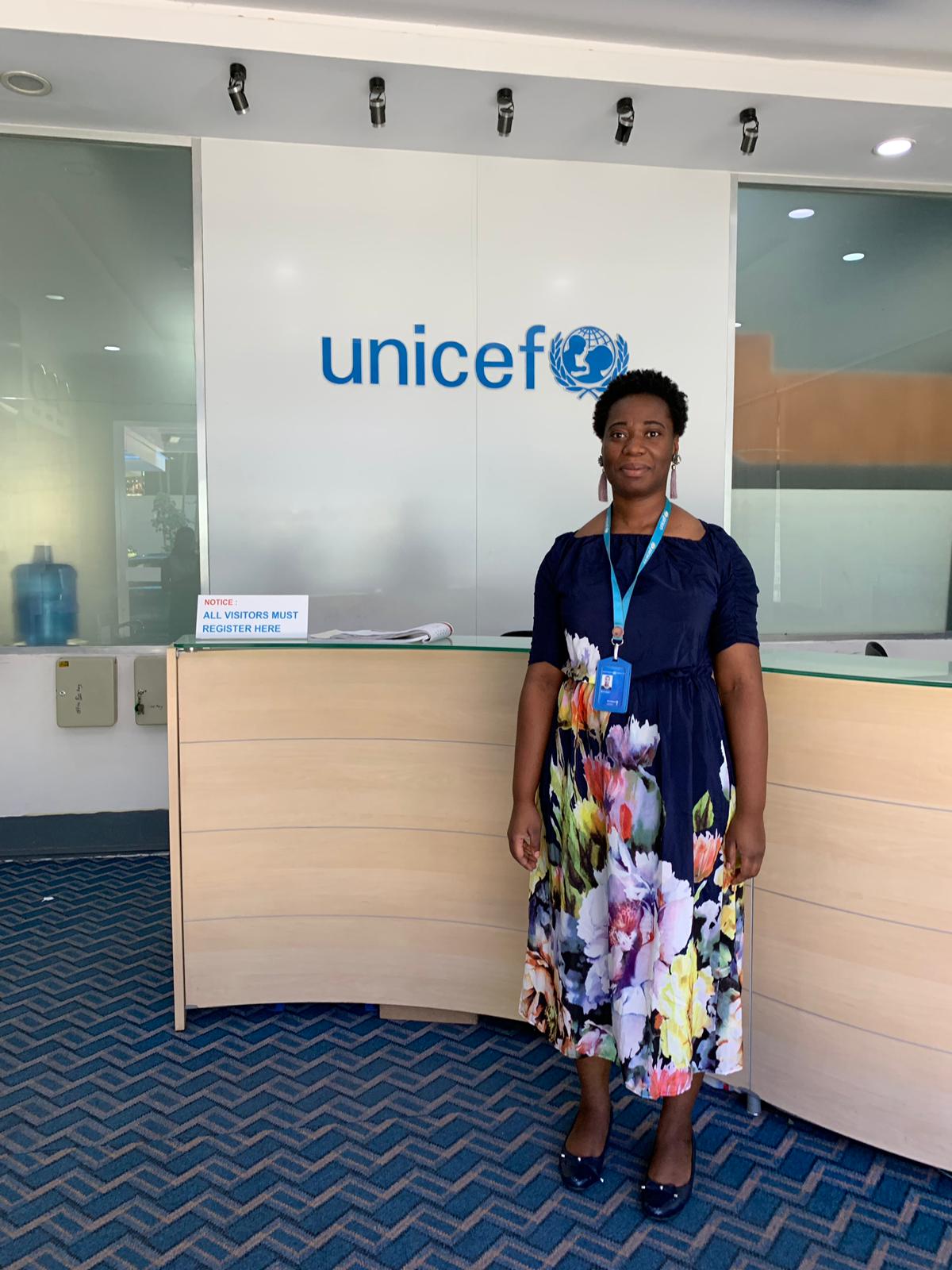 Marie Djeni standing in front of a reception desk and wall with the UNICEF logo.