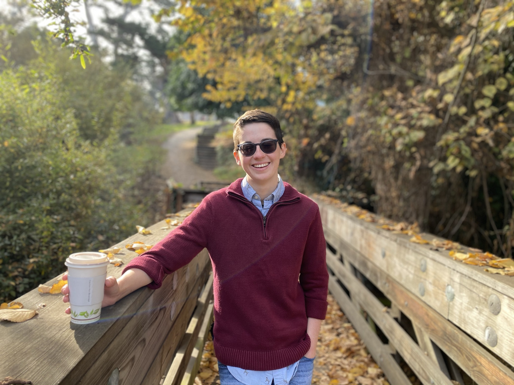 Student standing on a walking bridge, holding a coffee, wearing sunglasses and smiling