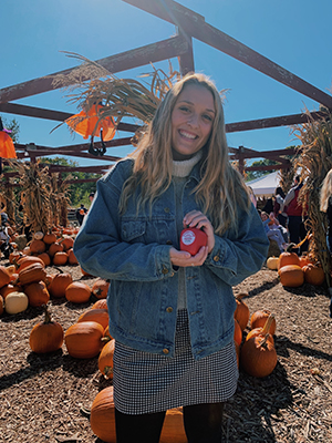 CBE Summer Fellow 2021-Elise Birkett-fall scene at pumpkin patch, elise holds her little pumpkin, smiling, head tilted, long blond hair, wearing jean jacket, b/w skirt with black tights