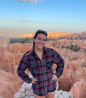 CBE Summer Fellow 2021-Grace Hansen-wearing flannel shirt and shorts, hands on hips, leaning right, smiling against the stunning background of Bryce Canyon National Park, Utah, pink spires, blue skies