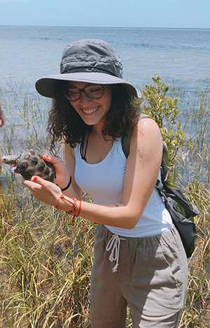CBE Summer Fellow 2021-Laura Sanchez-in a sun hat, shorts, white tank top standing in a marsh, holding a turtle, smiling
