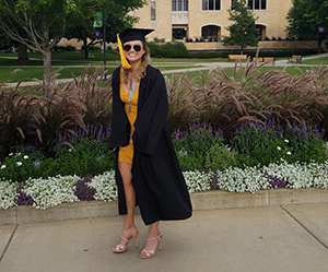 CBE Summer Fellow 2021-Natasha Lara Hood in her graduation garb, nice dress, black robe, square hat with tassle full body photo against a university campus