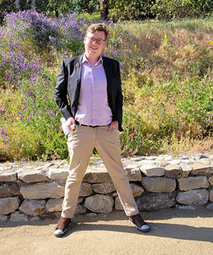 CBE Summer Fellow 2021-Sam Naujokas-standing with a wide stance, hands in pockets, smiling in the sunshine, kaki pants, button up shirt, blazer in front of a garden