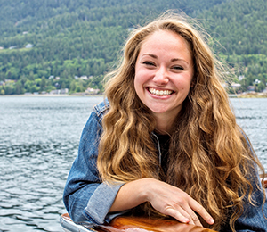 CBE Summer Fellow 2021-Olivia Cable-Barber--smiling brightly with long blondish hair, hands crossed in front, against background of lake or body of water, green trees or land behind