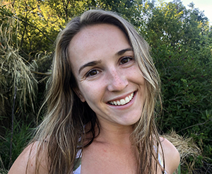 CBE Summer Fellow 2021-Mary Patenburg-headshot that is casual, her head tilted, smiling, bare shoulders, sandy hair to shoulders, looking happy and windswept against a lush green shrub background