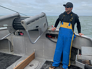 CBE Summer Fellow 2021-Nick Rahaim-wearing blue and yellow rubber overalls on deck of Alaskan fishing vessle where he leans against the rail, with sunglasses and ball cap, cold pale blue ocean behind him