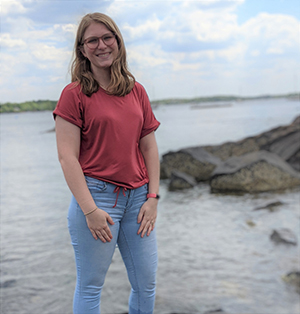 CBE Summer Fellow 2021 Tabitha Birdwell standing in front of a bay or lake, gray water, brown rocks, green land and blue sky with big white cloud behind her, she stands with hands on thighs, smiling, reddish shirt and blue jeans, wears glasses with shoulder length light brown/blondish hair