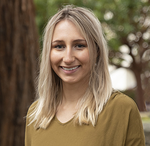 CBE Summer Fellow 2021-Diane Sikkens-wearing a professional looking mustard colored blouse, smiling brightly looking straight at the camera, with shoulder length blond hair, a beautiful redwood tree trunk and forrest behind her