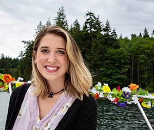 Heather Cook smiling with big green eyes, short hair, leaning against the railing of a sailboat that is decorated with a gardland of colorful flowers, with gray waters of a bay, and conifers and big green trees in the background