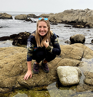CBE Summer Fellow 2021-Emily Stokes-she is crouching down on the buff colored granite rocks of a tidepool, with gray ocean waters and brown kelp seen behind her; she is smiling smartly with one hand on the rocks, and sunglasses holding back her hair