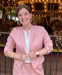 Madeline Warner in a CUTE pink business suit, smiling with bright eyes, standing in front of an antique merry-go-round that is lit up and pretty