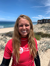 Madison Springfield smiling joyfully, in a wetsuit and red swim shirt with the logo for the Wahine Project, the beach, ocean, and blue sky behind her