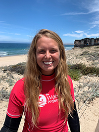 Madison Springfield smiling joyfully, in a wetsuit and red swim shirt with the logo for the Wahine Project, the beach, ocean, and blue sky behind her 