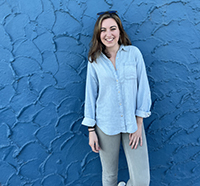 Molly Ryan standing before a bright blue stucco wall, in a blue blouse, looking sharp with her dark hair, and bright smile