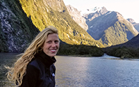 Liz Hofius looking gorgeous, long blond hair blowing in wind, stading in front of an alpine lake, snow capped mountains behind