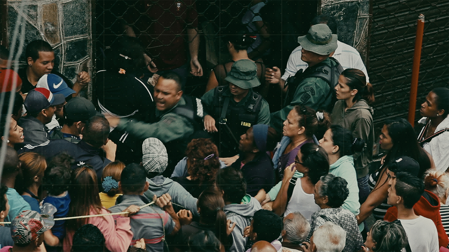 A scene of a crush of people, upset, yelling, some holding a rope line--its a food panic in Venezuela, year unknown