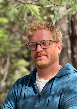 Investigative reporter Nate Halverson in a sunlit forest, strawberry blond/red hair and beard, glasses, smiling slightl