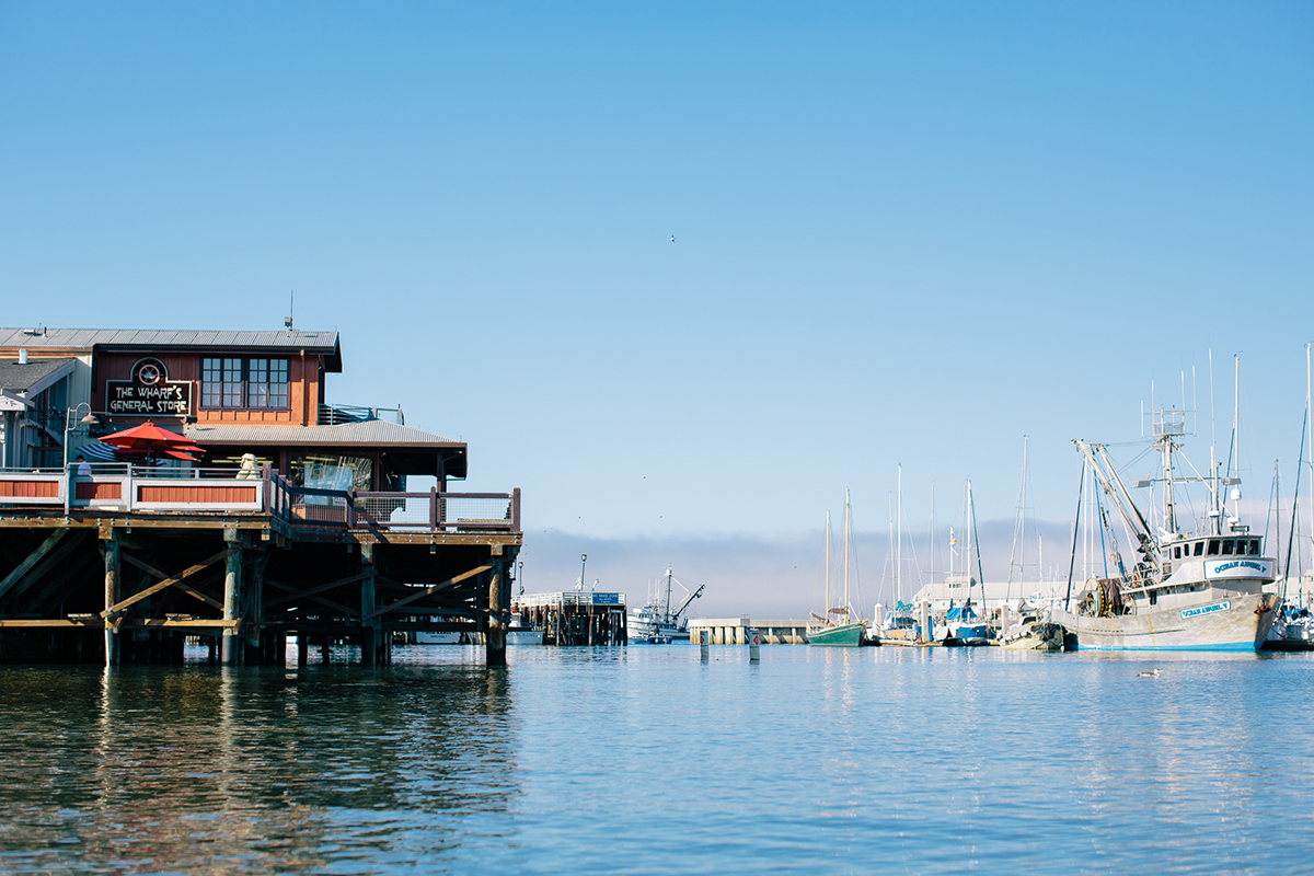 The "tourist wharf" in Monterey CA, a restaurant over pilings above a calm harbor, with commercial fishing boats and recreational sailboats in the background, blue sky, blue water, brown wharf, pink-orange restaruant, and white boats. 