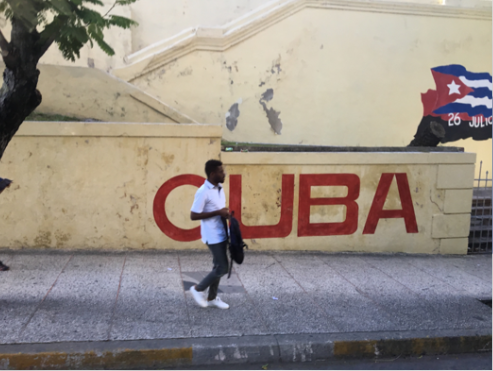 African American man walking on the street in front of a mural on a wall which reads "Cuba" in red letters