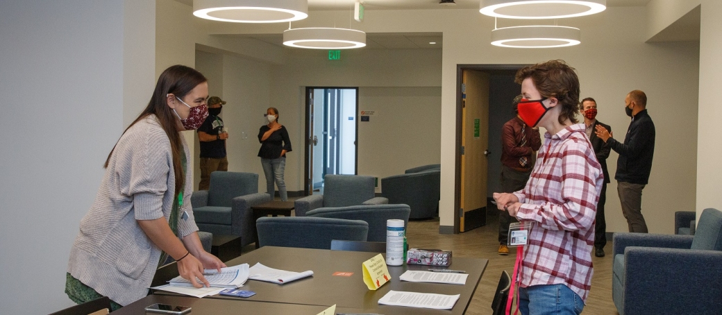 Woman wearing masks stands across a table from a younger person wearing a mask