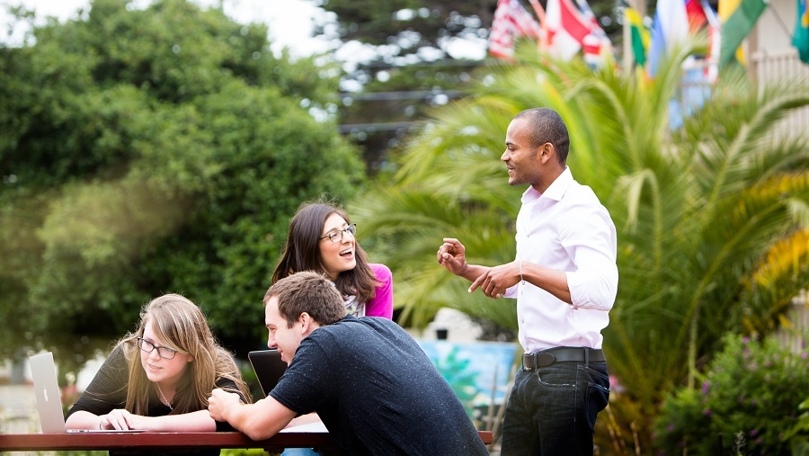 Group of four students around a table outside looking at a laptop.  Palm trees and flags in the background.