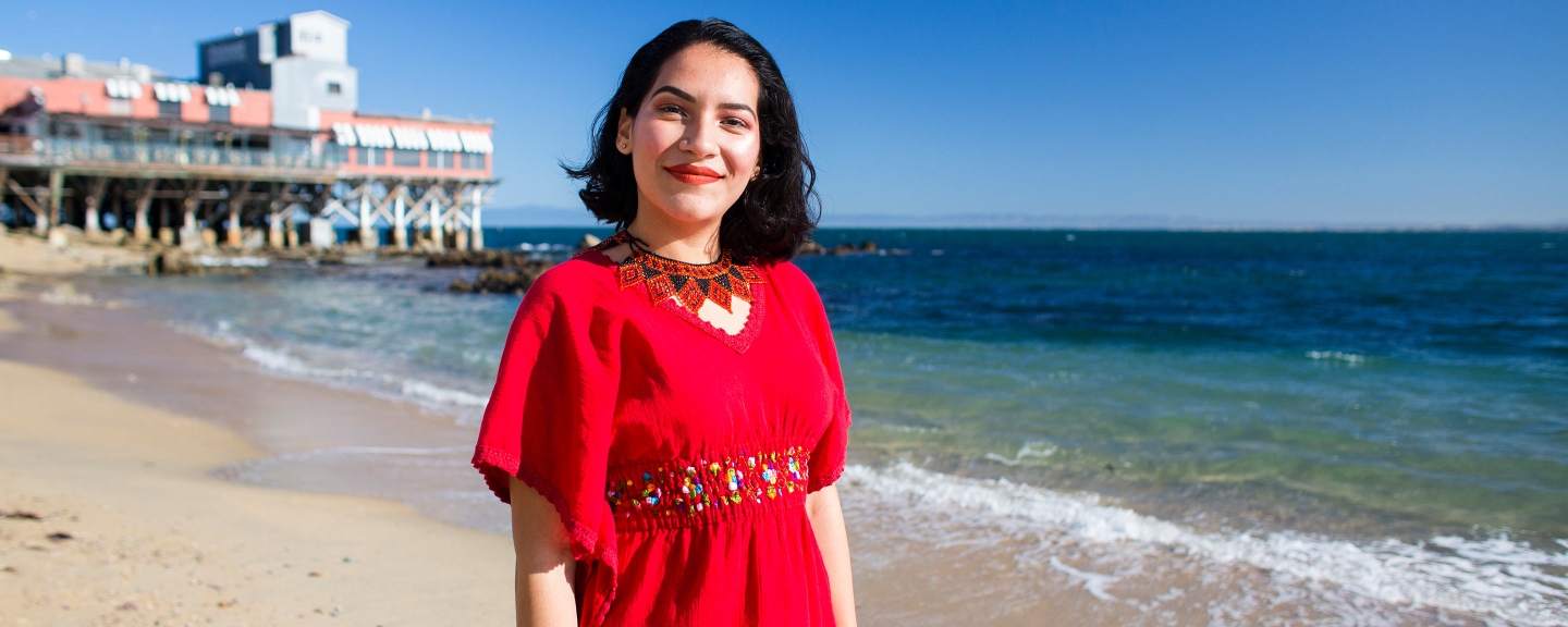A student stands on the beach with the pier and Monterey Bay behind her.