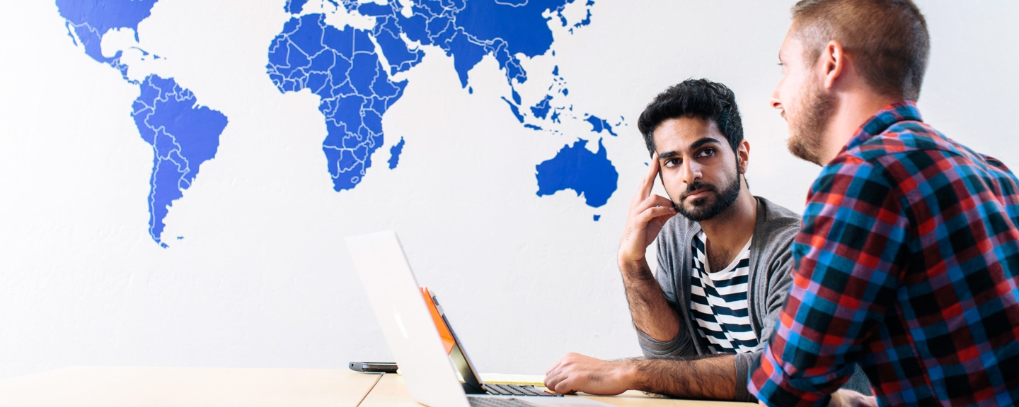 Two students at computers are discussing their work in front of a world map.