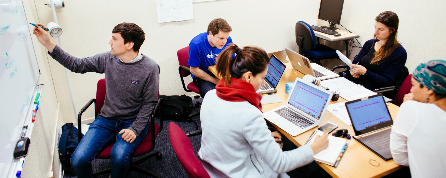 Students working together in a classroom setting with computers and white board.