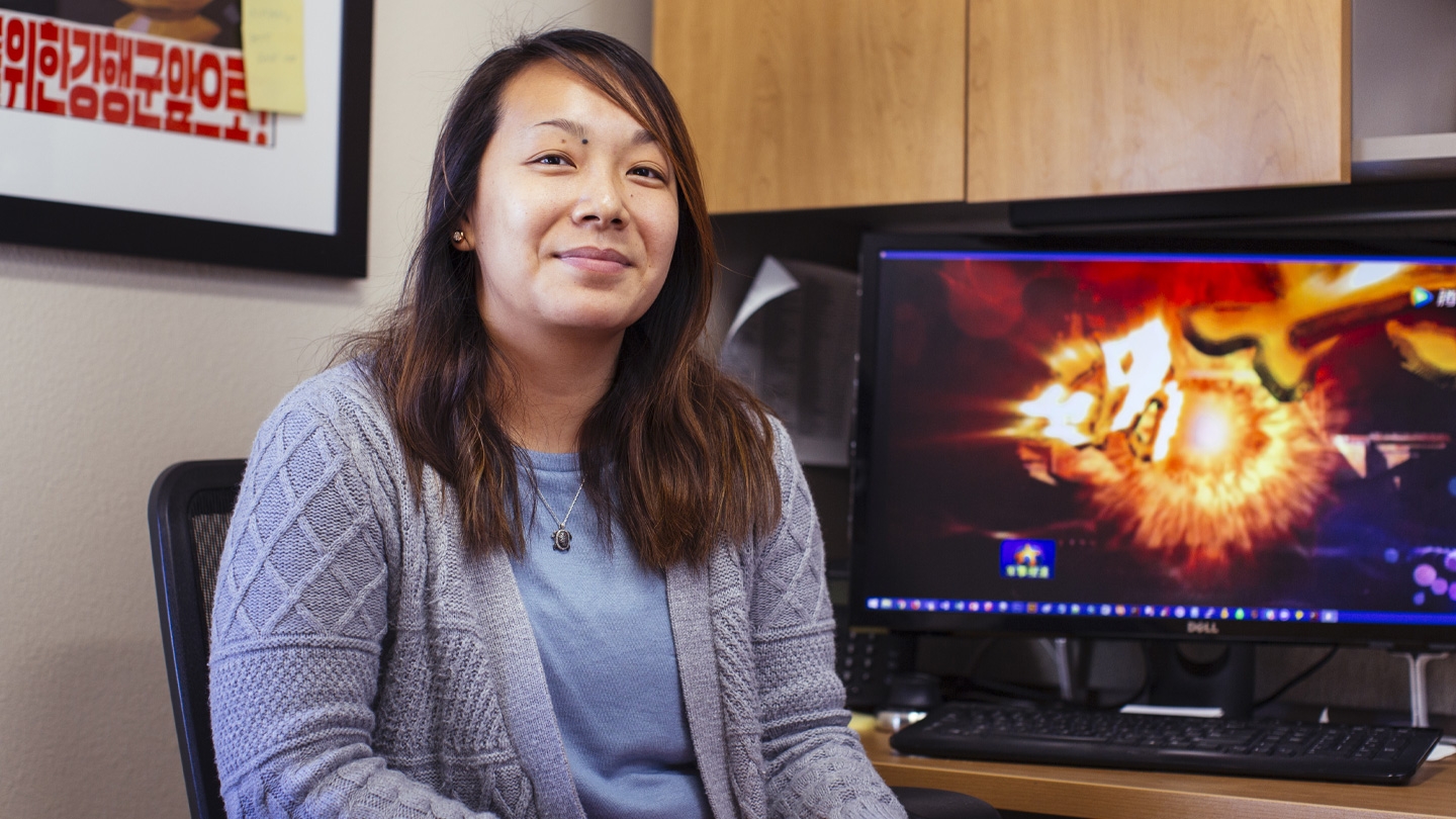 Student, Olivia Hallam, sitting and smiling at the camera, her work computer and a poster with korean letters are behind her.