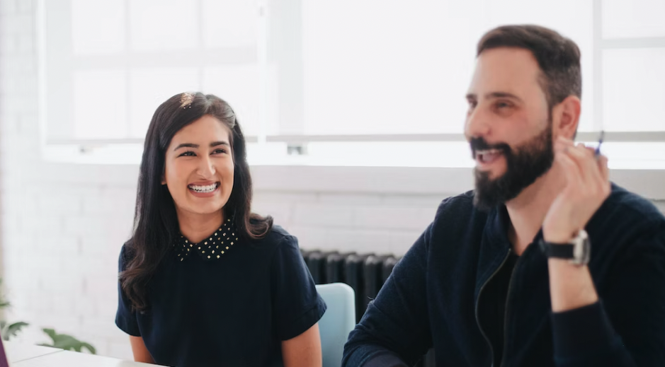 one woman and one man sitting at a desk laughing and looking at each other, one is looking away