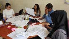 Four people sitting around a table looking through papers and working on their development project. There is one female to the far left in a white top with brunette hair pulled up into a ponytail. The right of her there is another female holding up a paper reading it. Second to the right, a man is sitting with a pencil in his hand adjusting his glasses. On the far right a woman wearing hijab is looking at a list of rules.