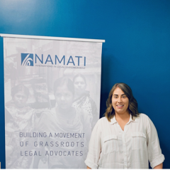 A girl with brown hair in a white shirt standing in front of a sign for Namati with a blue wall as the background.