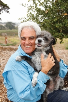 Picture of the Mr. Bruce Delgado, Mayor of the City of Marina, shown smiling, with white hair and in a blue shirt, holding a CUTE doggie who is black with a white belly and white face, both in a beautiful oak woodland