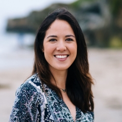 Ms. Mariko Powers, headshot, with a bright smile, dark, long hair, smiling eyes outdoors with a beach and cliff in the background