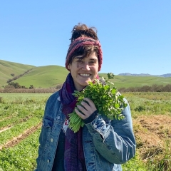 Ms. Ellen Olack smiling brightly with a bright green bunch of cilantro in her hand, standing a farm field with rows of green crops, and soft green rolling hills in the background, bright blue sky.  She is wearing a pink headwarmer, purple scarf, and blue jean jacket.  