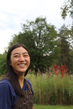 A young woman in brown overalls and blue shirt, smiling brightly against a lush green background, grass, tall grass, and trees, a red sculpture is hidden in the grass