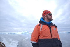 Ari S. Friedlaender, PhD, looking badazz in profile against an Arctic sky--he studies whales and is awesome. White/blue sky, white/blue glacial ice and ocean water, he is wearing an orange/black jacket, red snug cap, and sunglasses.