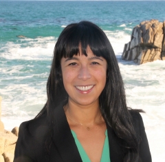 Angelina Skowronski smiling brightly, with bright eyes, long black hair with bangs, against a background of the light blue ocean and white waves crashing on brown gold rocks