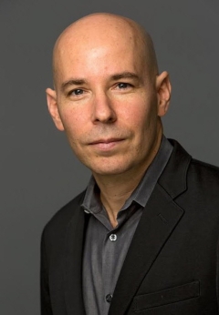 Raz Godelnik, headshot of a serious and smart looking man, bald, wearing a black business blazer over a grey collared shirt.