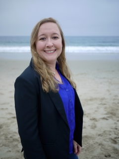 Amanda Leinberger, smiling brightly, standing on a beach with a blue ocean and white waves behind her, looking professional and cheerful