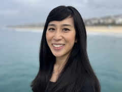 Author and Journalist Rosanna Xia looking at the camera with a bright and penetrating gaze, warm smile, with long black hair against a light blue sea and strip of coastline in the background