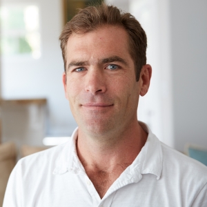 Peter Macy headshot, white polo shirt in a home setting, smiling a sly and intelligent smile