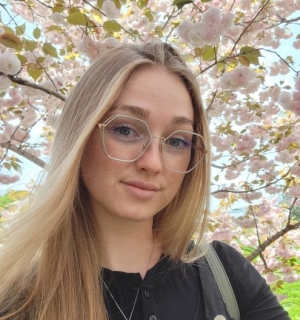 photo of smiling Dayna Brown; hexagonal wire-rimmed glasses, blonde, cherry blossoms behind her