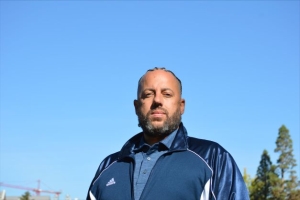 Jody Lykes photo of a man standing with blue sky behind him wearing a blue track jacket