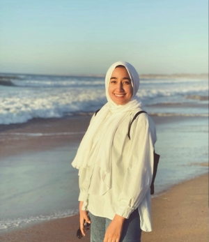 Photo of young woman, Marwa, standing by the Pacific ocean wearing a white, long-sleeved blouse and a white hijab head scarf.