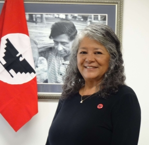 photo of latina woman, Teresa Romero, standing in front of a black and white image of a farm worker carrying farm poles over his shoulder.